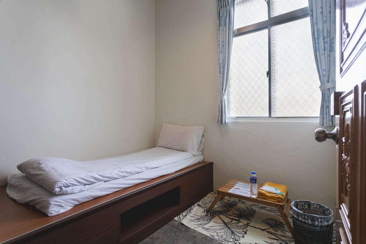 A small, single-bed room with a wooden bed frame, a window with curtains, a small table with a bottle and a folded towel, and a trash bin.