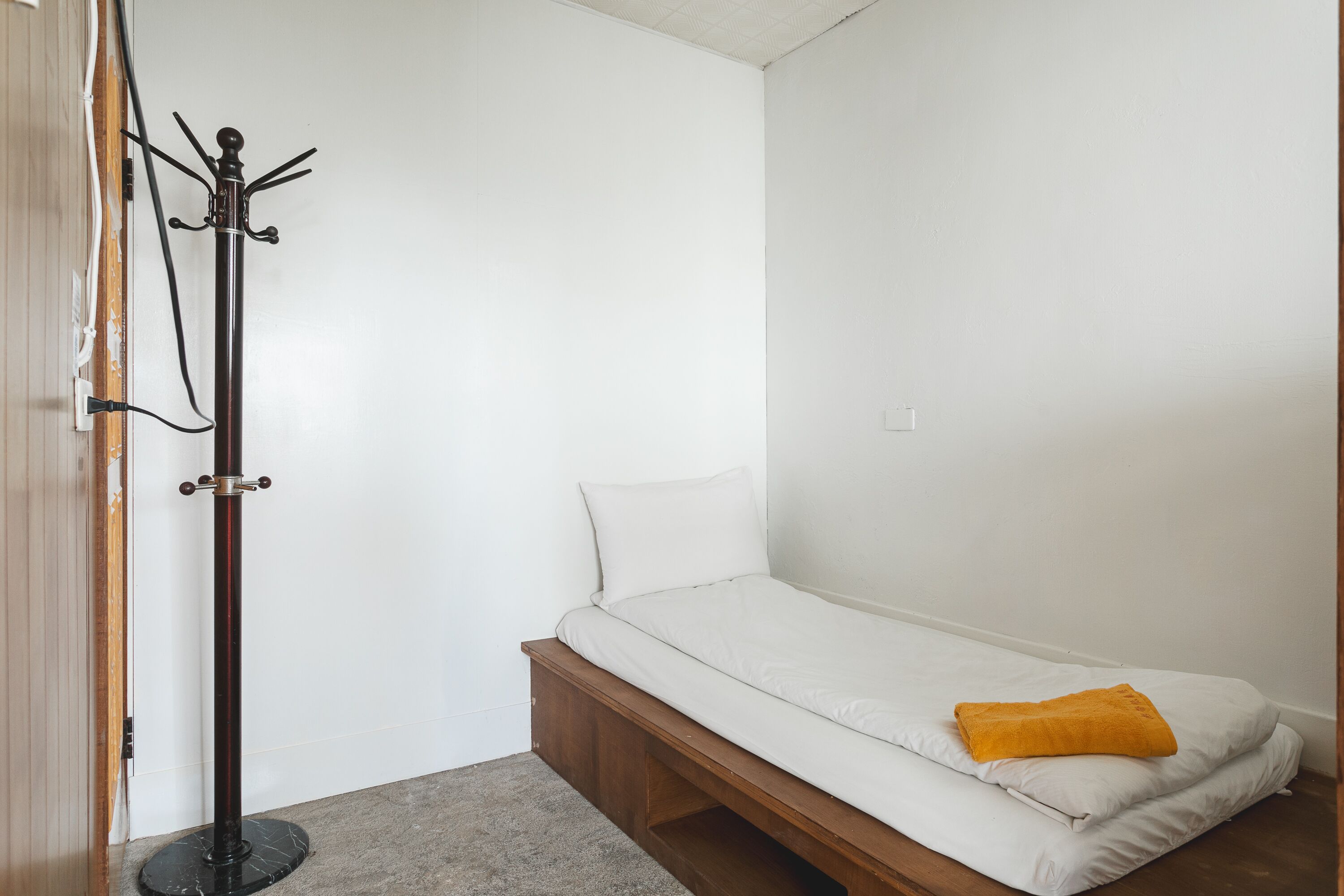 A small, single-bed room with a wooden platform bed, a coat rack, and a grey carpet.