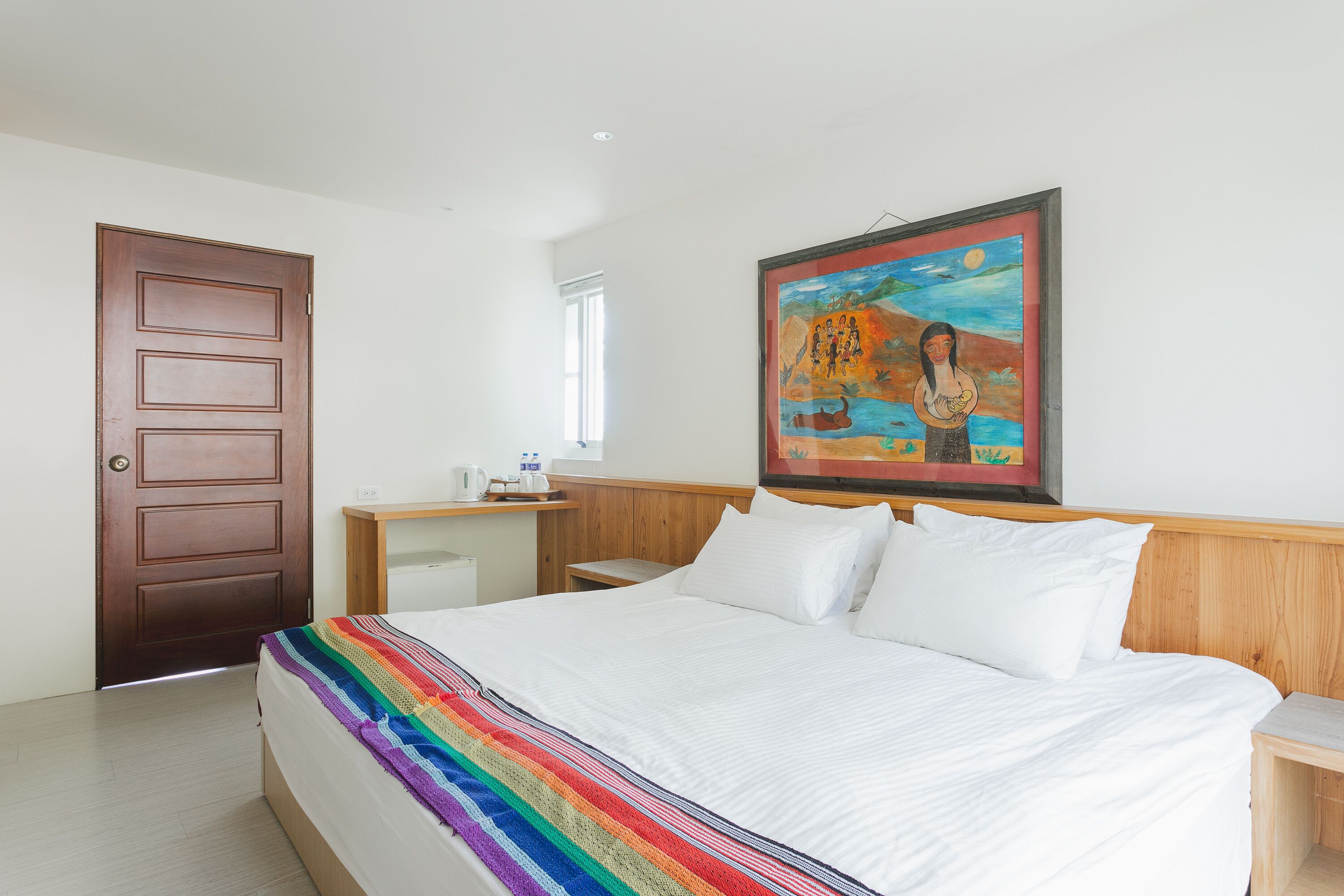 Une chambre d’hôtel avec un lit, une tête de lit en bois, une table de chevet, un tableau au mur et une porte.