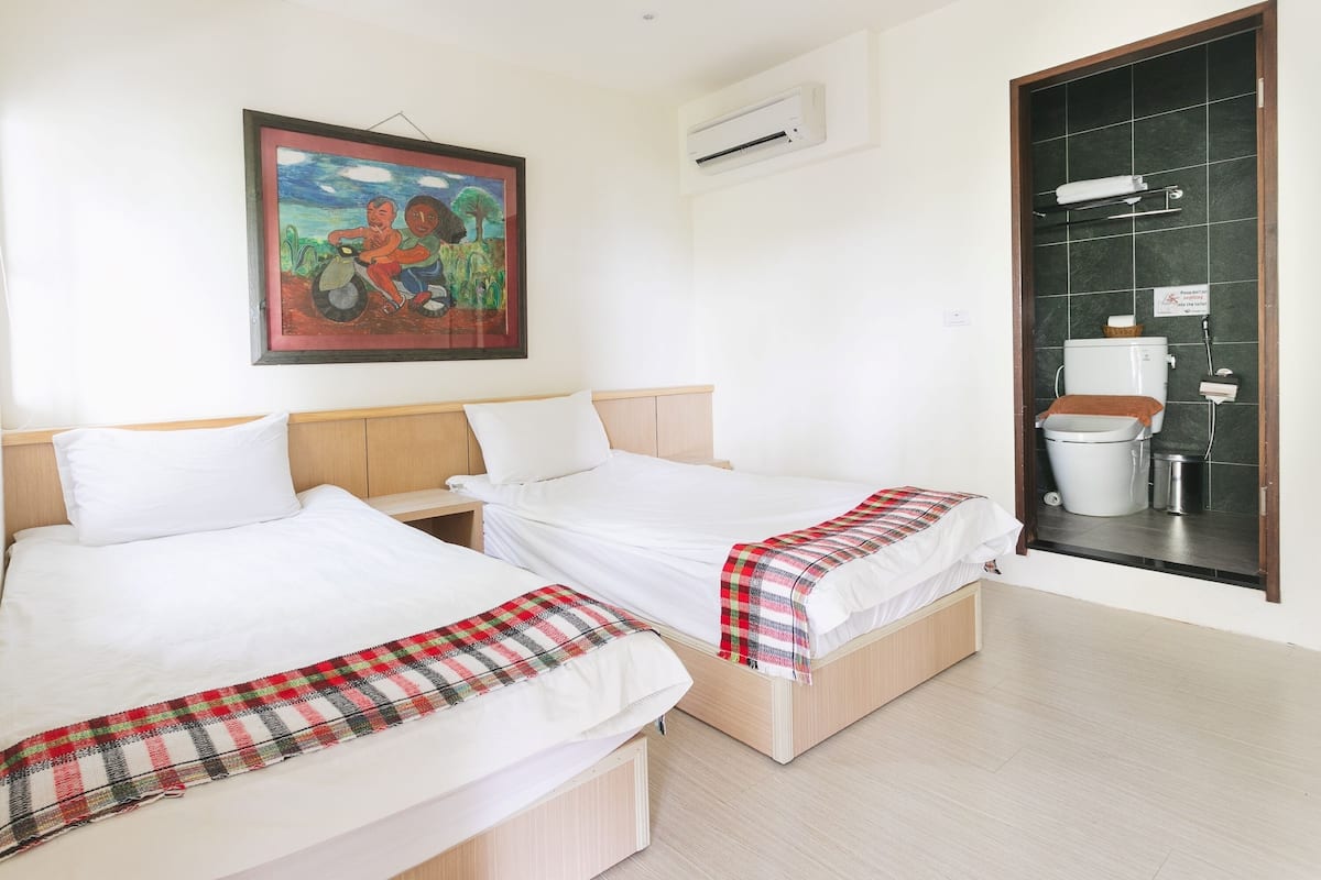 A hotel room with two beds, a painting on the wall, an air conditioning unit, and a bathroom with a toilet and sink.
