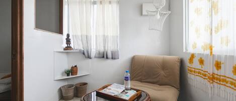 A room with a sofa, a small table with a tray, a water bottle, a wall-mounted shelf with decorative items, and a window with curtains.