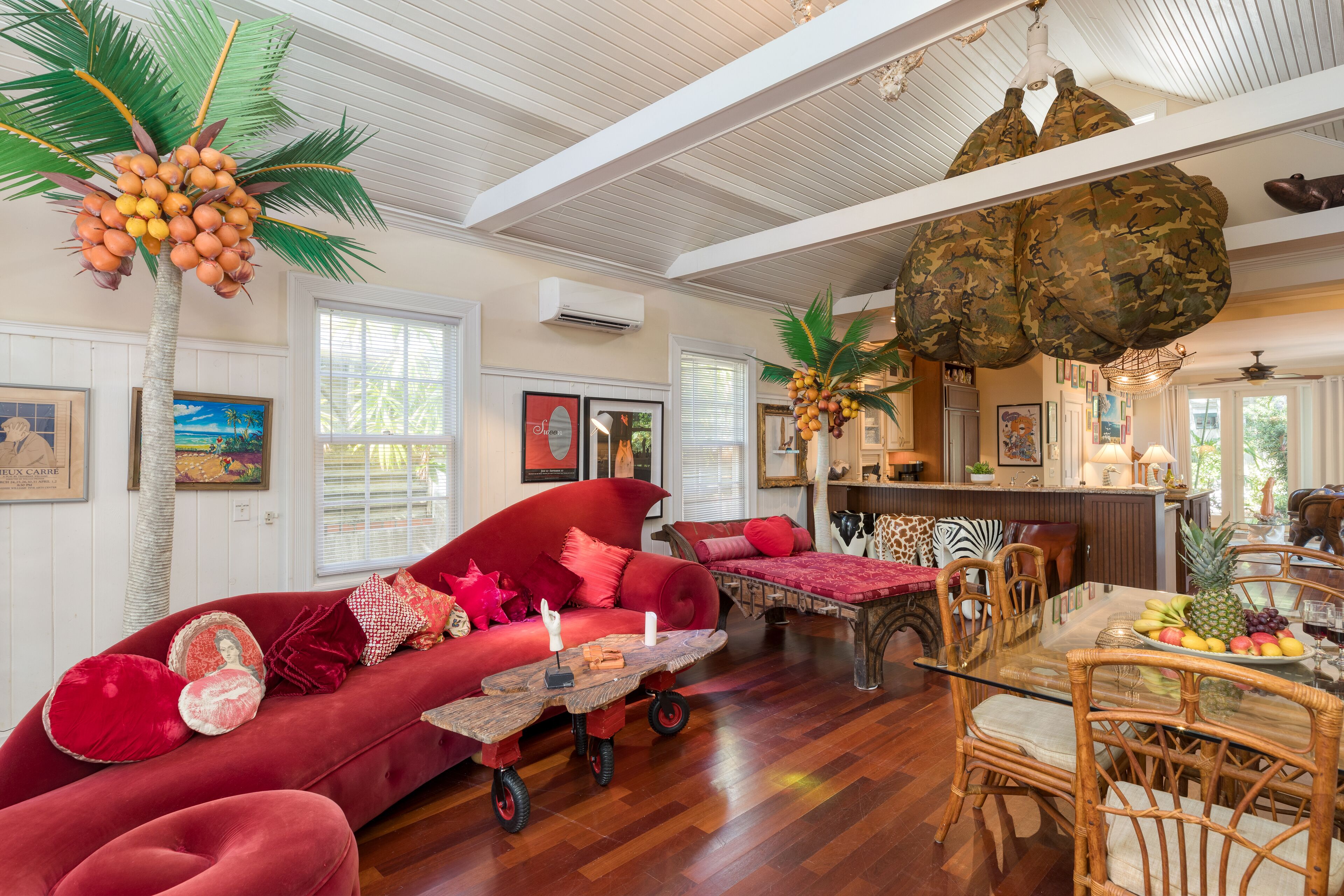 A spacious living room with a red sofa, wooden dining table, and tropical decor including a palm tree and a hanging coconuts.
