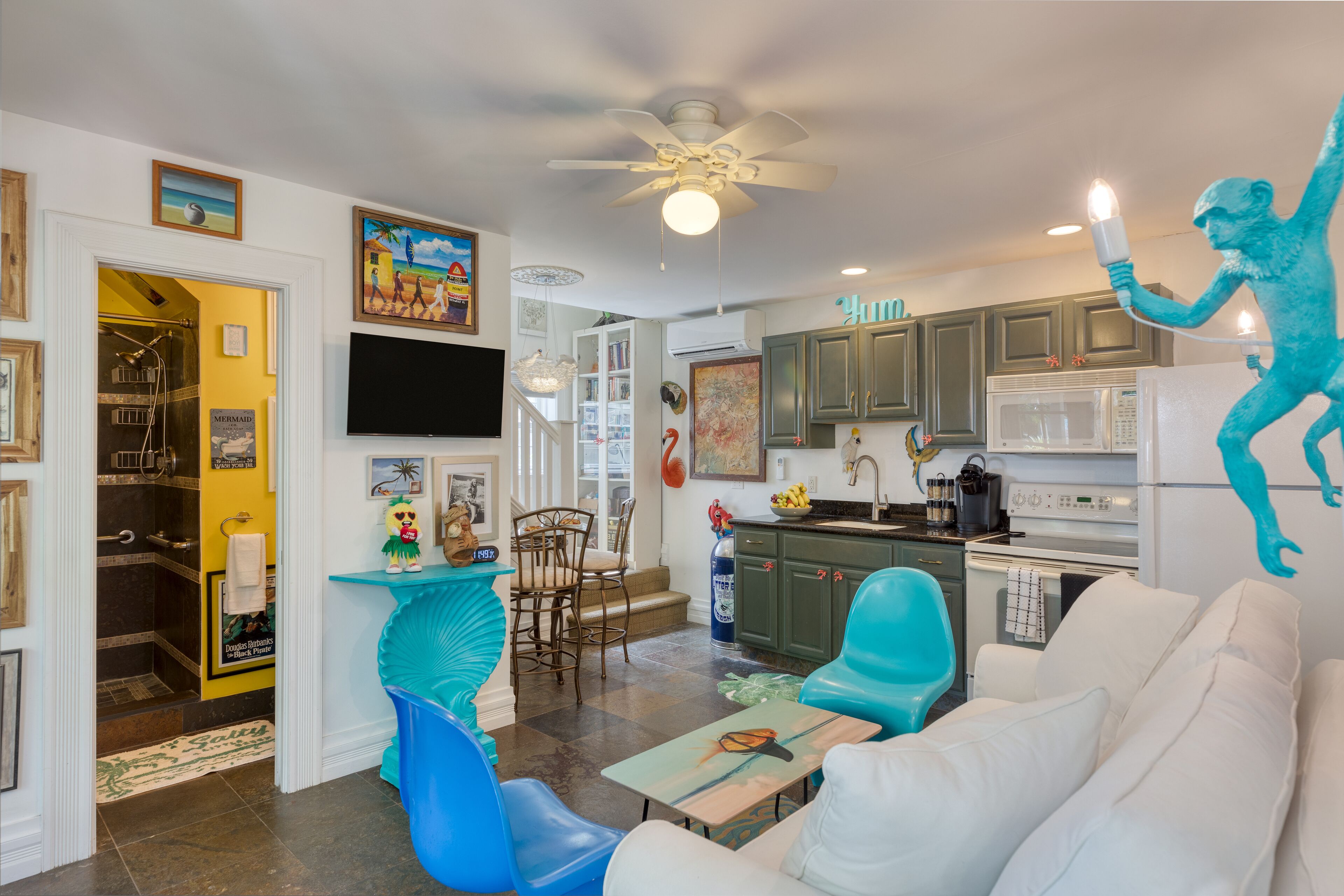 Un salon moderne avec un canapé blanc, des fauteuils turquoise et un ventilateur de plafond. On y trouve également un coin cuisine avec des meubles verts et un four à micro-ondes.