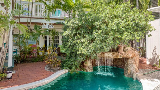 Une piscine agrémentée d’une cascade, entourée d’une végétation luxuriante, et un bâtiment blanc doté de balcons.