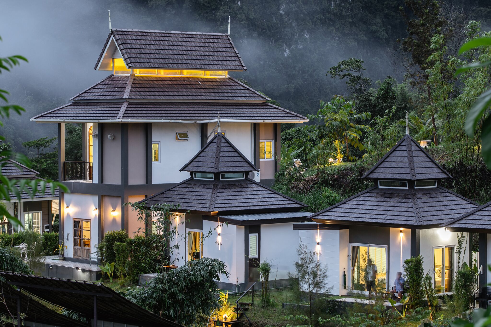 Uma casa moderna com um edifício central amplo e duas estruturas menores anexas, rodeada por vegetação exuberante e um cenário montanhoso.