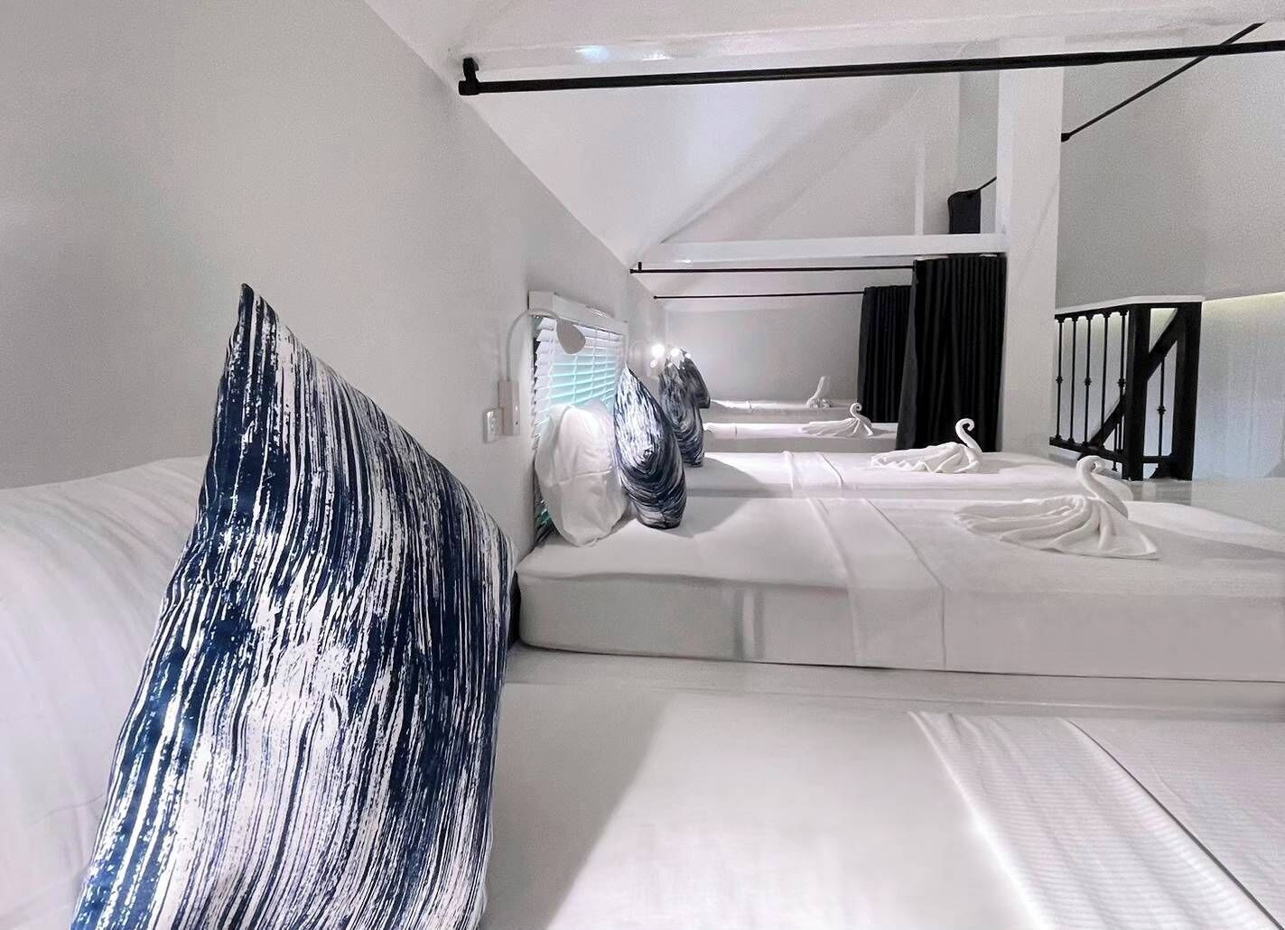 A modern hotel room with a minimalist design, featuring a large bed with a white and blue patterned headboard, a separate sleeping area with a white bed, and a black railing along the window.