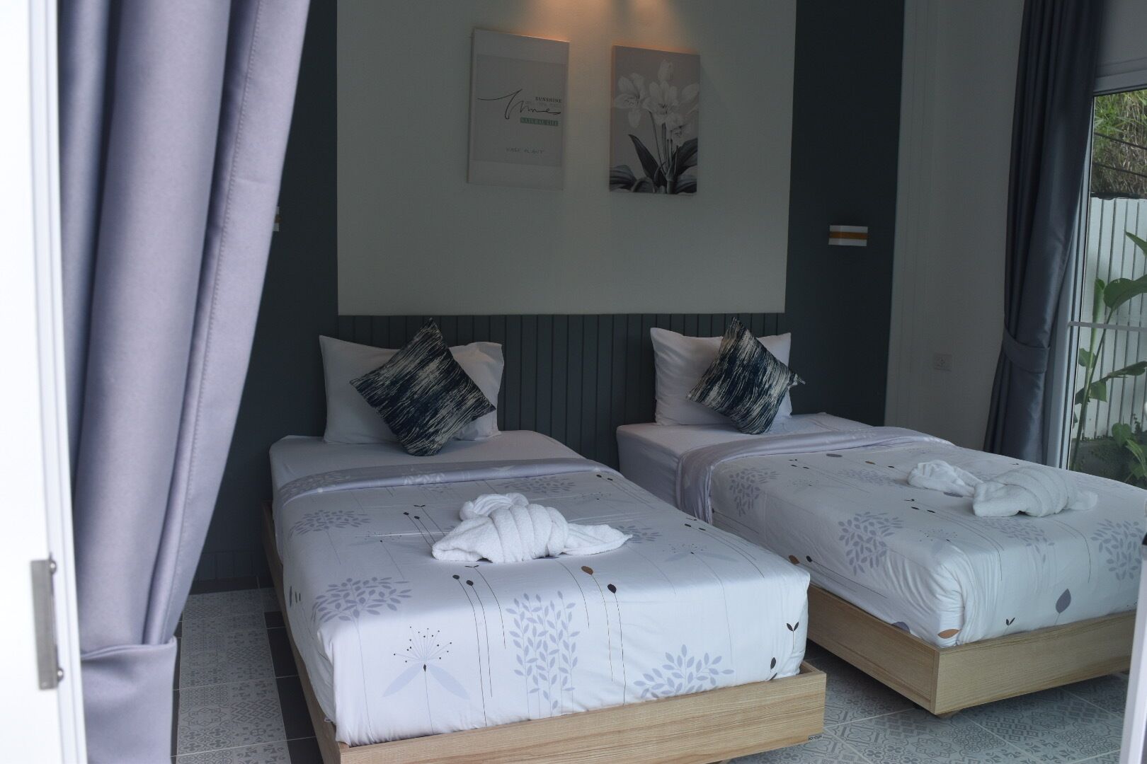 A hotel room with two beds, each with a white bedspread and a single black and white pillow. There is a framed artwork above the beds and a potted plant to the right.