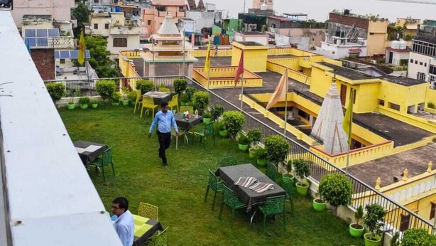 A rooftop terrace with green chairs and tables, a view of the cityscape, and a person walking. - Hotel Banaras Haveli (Varanasi)