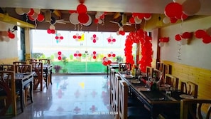 Guests can dine in a spacious room with wooden tables and chairs, a glass door leading to an outdoor area, and a high ceiling with visible beams.