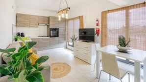Une cuisine moderne avec des meubles en bois, une cuisinière à gaz et un four encastré. On y trouve un coin repas avec une table et des chaises blanches, ainsi qu’un téléviseur posé sur un meuble.