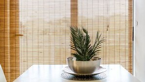 Un coin repas avec une table blanche, un bol contenant une plante et une fenêtre avec des stores.
