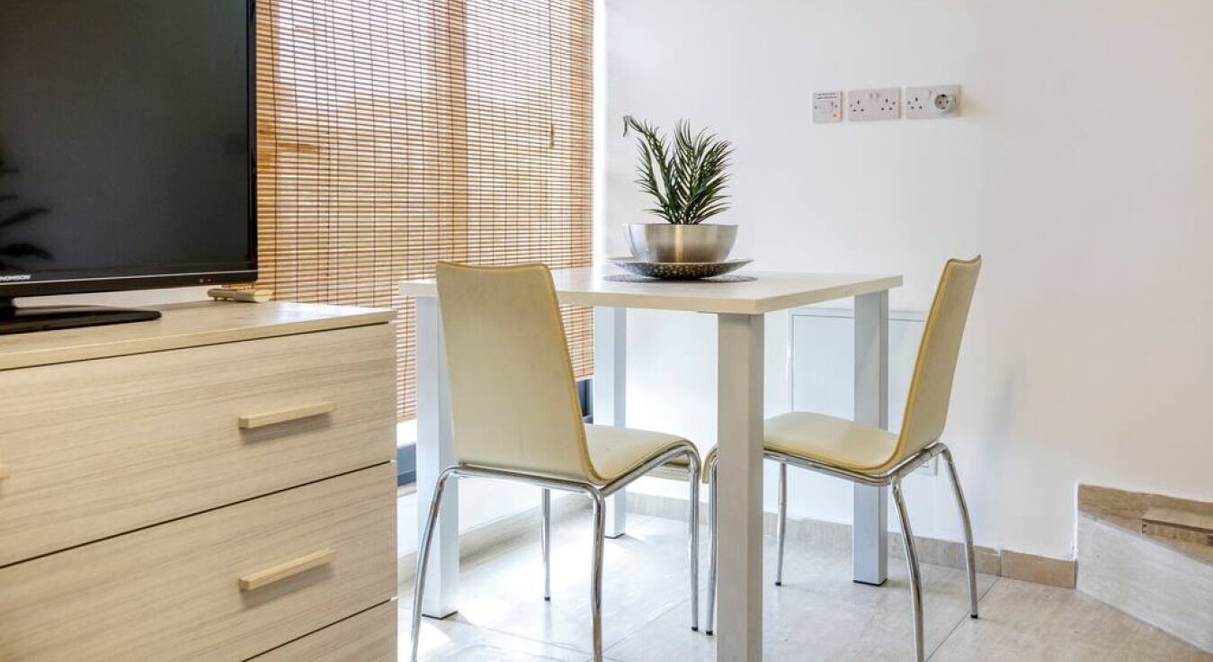 A modern room with a wooden dresser, a flat-screen TV, two chairs, a small table with a potted plant, and a window with blinds.