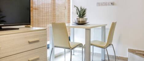 A modern room with a wooden dresser, a flat-screen TV, two chairs, a small table with a potted plant, and a window with blinds.