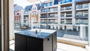 Un balcón con una mesa de vidrio y mobiliario de mimbre, con vista a una calle con edificios modernos y tradicionales.