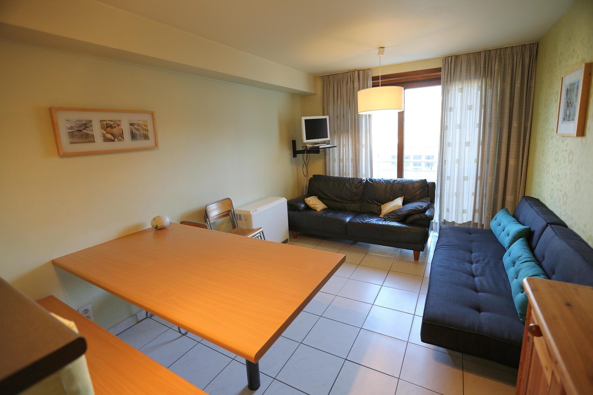 A room with a wooden table, a black sofa, a television on a stand, and a window with curtains.