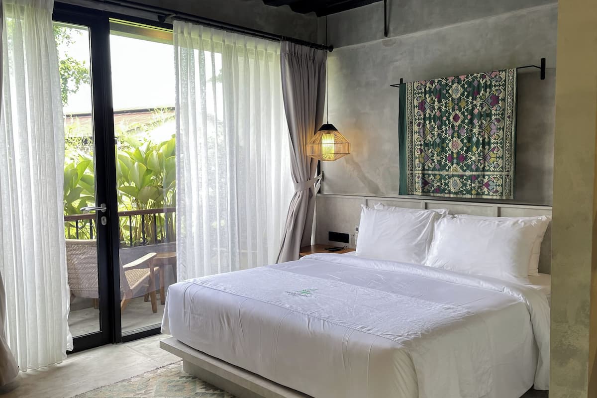 A bedroom with a bed, a nightstand, a lamp, a view of greenery outside, and a patterned tapestry on the wall.