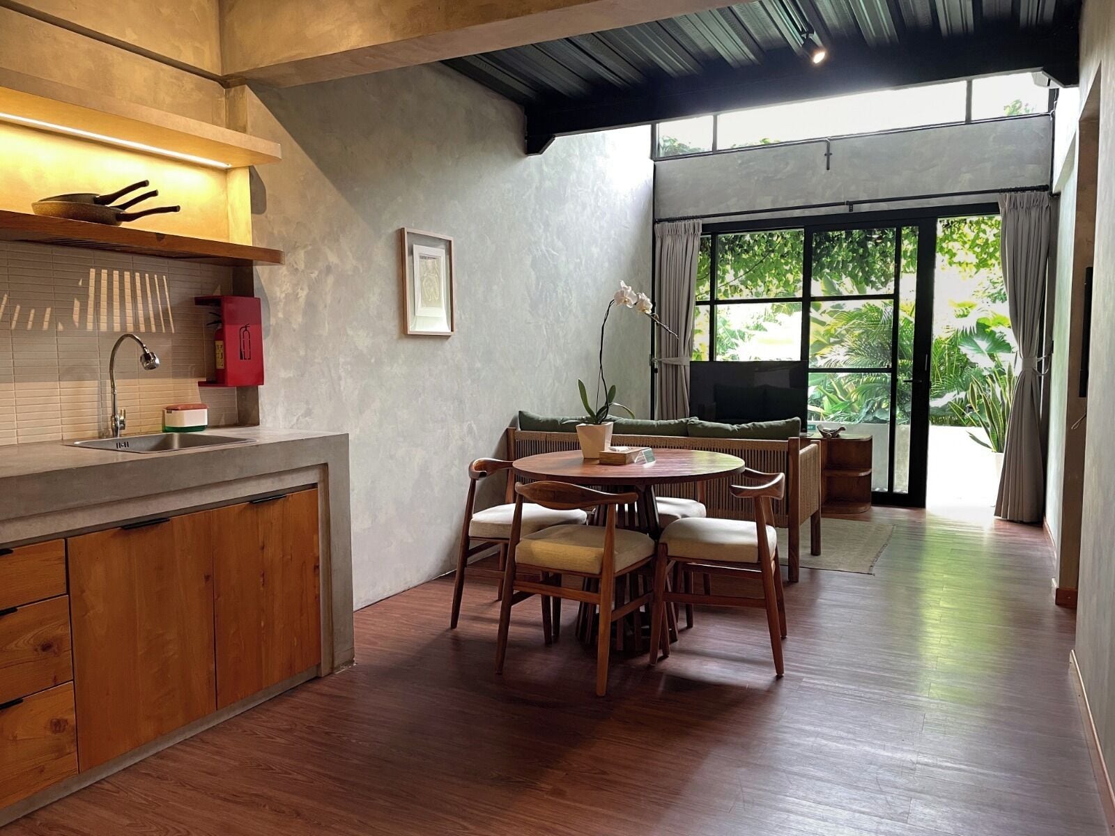 A modern kitchen with wooden cabinets, a dining area with a round table and chairs, and a large window with a view of greenery.