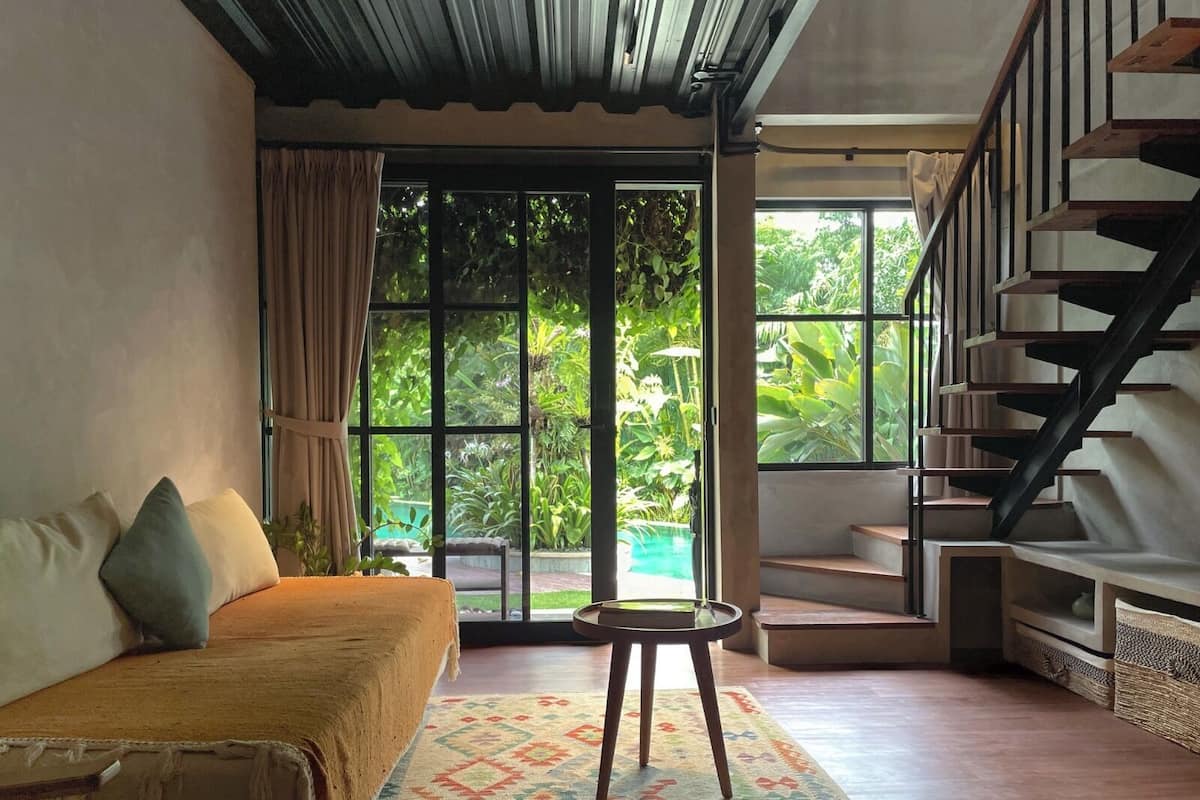 A room with a sofa, a small table, a staircase, and large windows with a view of greenery.