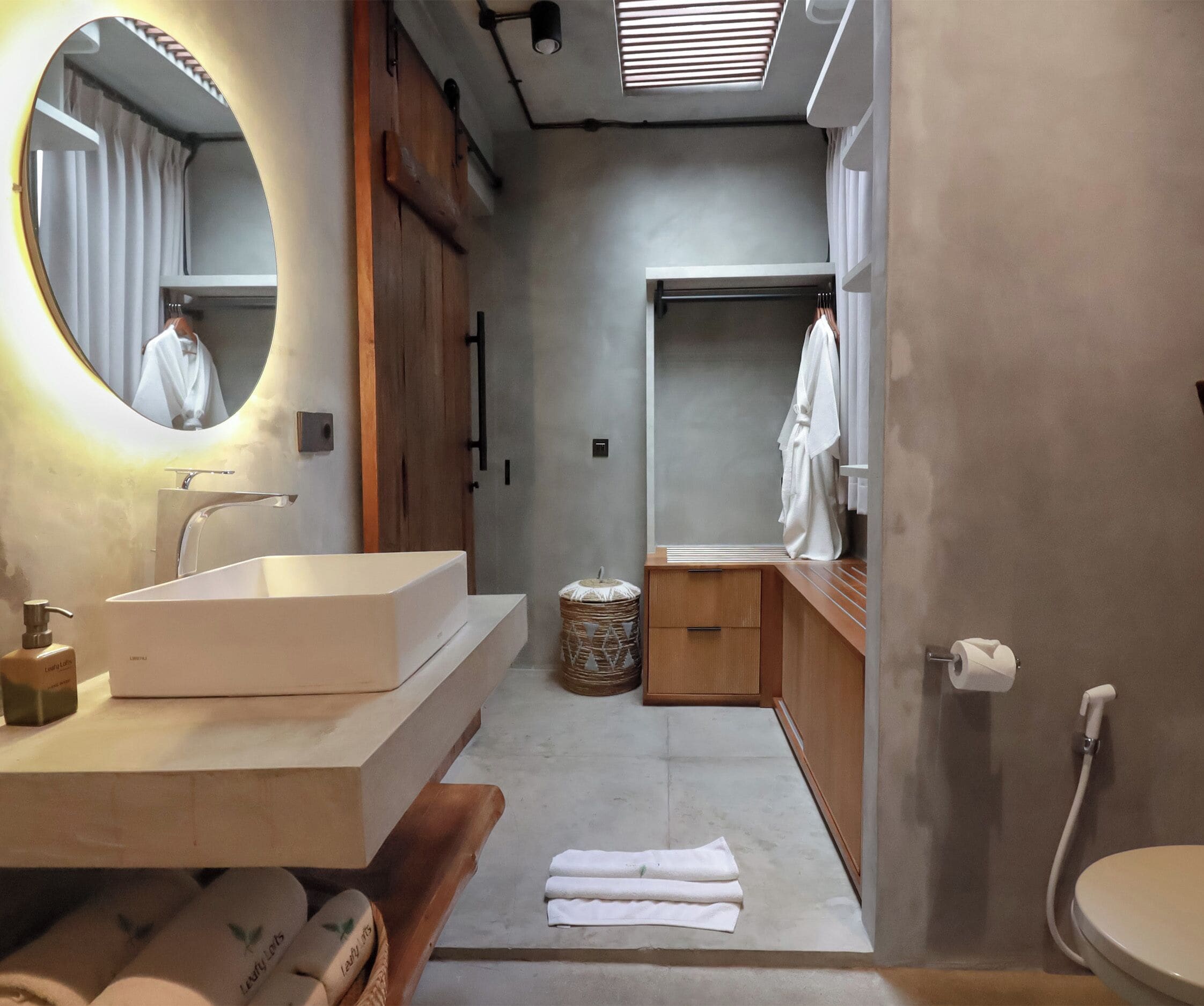 A modern bathroom with a large mirror, a sink, and a toilet. There is a wooden bench and a basket in the corner.