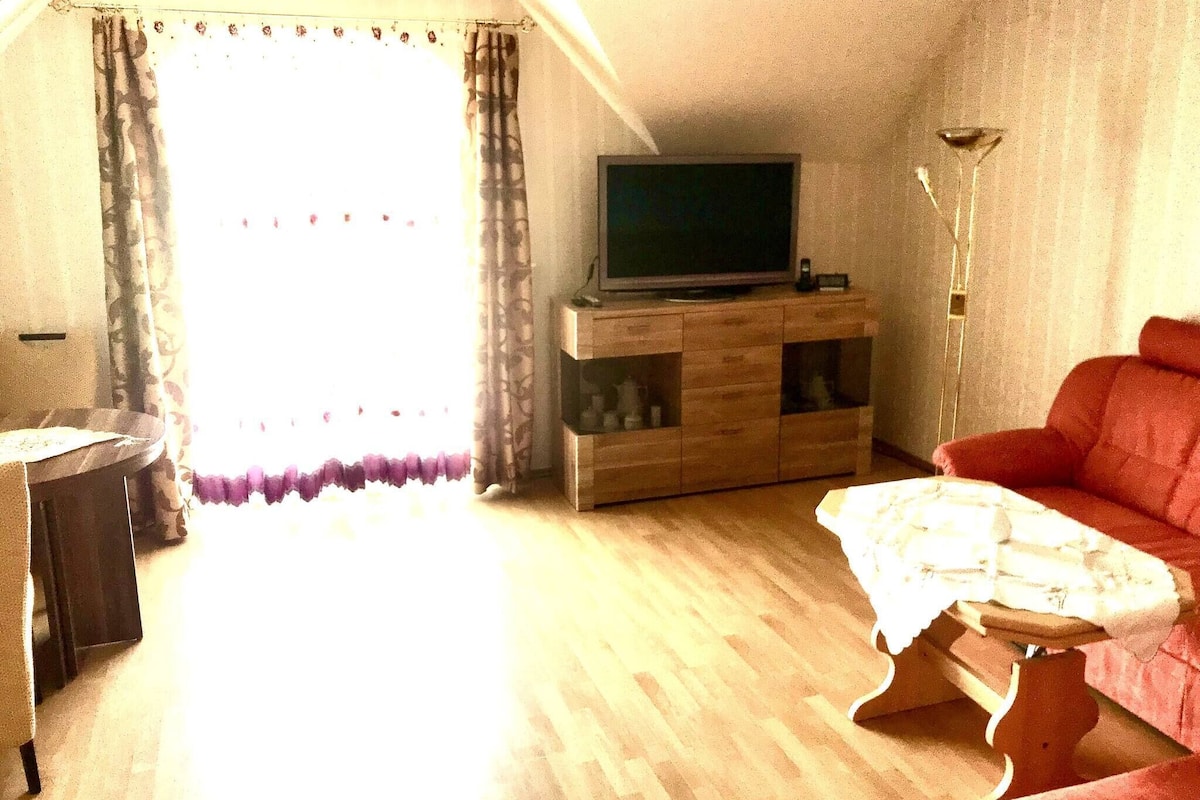 Un salon avec un téléviseur posé sur un meuble en bois, un canapé rouge et une table recouverte d’une nappe blanche.