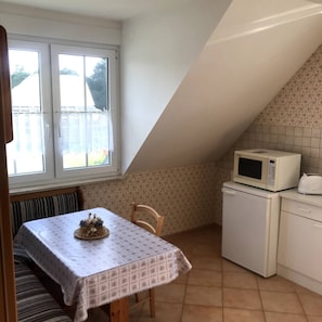 A small kitchen with a microwave, a refrigerator, a table with a placemat, and a window with curtains. - Room-haus Im Grünen House in the Green (Neuzelle)