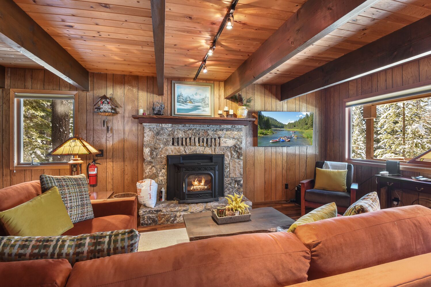 A cozy cabin interior with a stone fireplace, wooden walls, and a large window overlooking a forest.