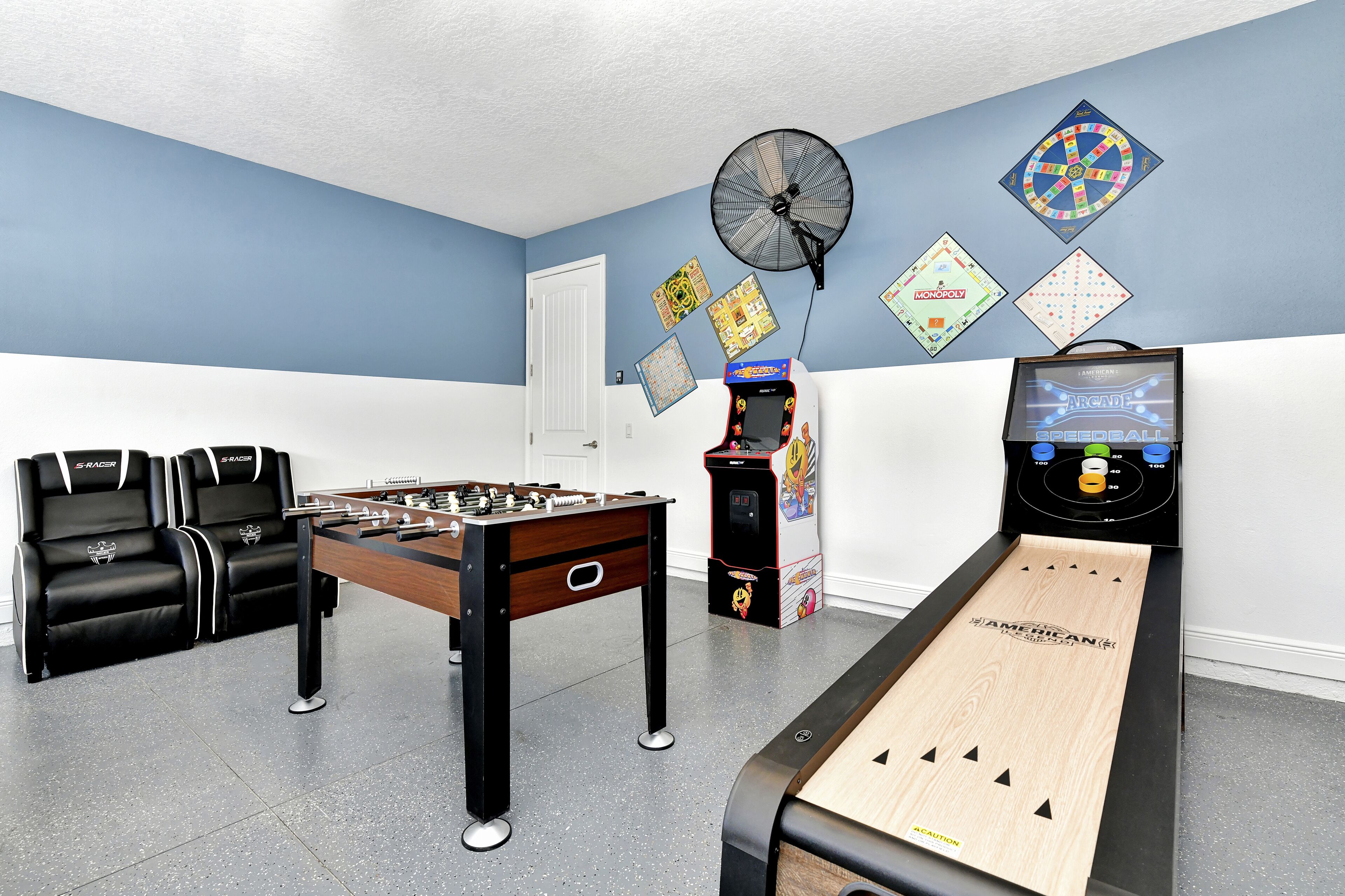 A room with a foosball table, a table tennis table, and a shuffleboard table. There are two sofas and a fan on the wall.