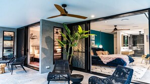 A modern interior with a large sliding glass door leading to a bedroom, a dining area with chairs and a table, and a ceiling fan.
