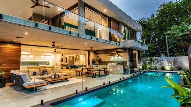 A modern two-story house with a swimming pool, outdoor seating, and a kitchen visible through large glass doors.