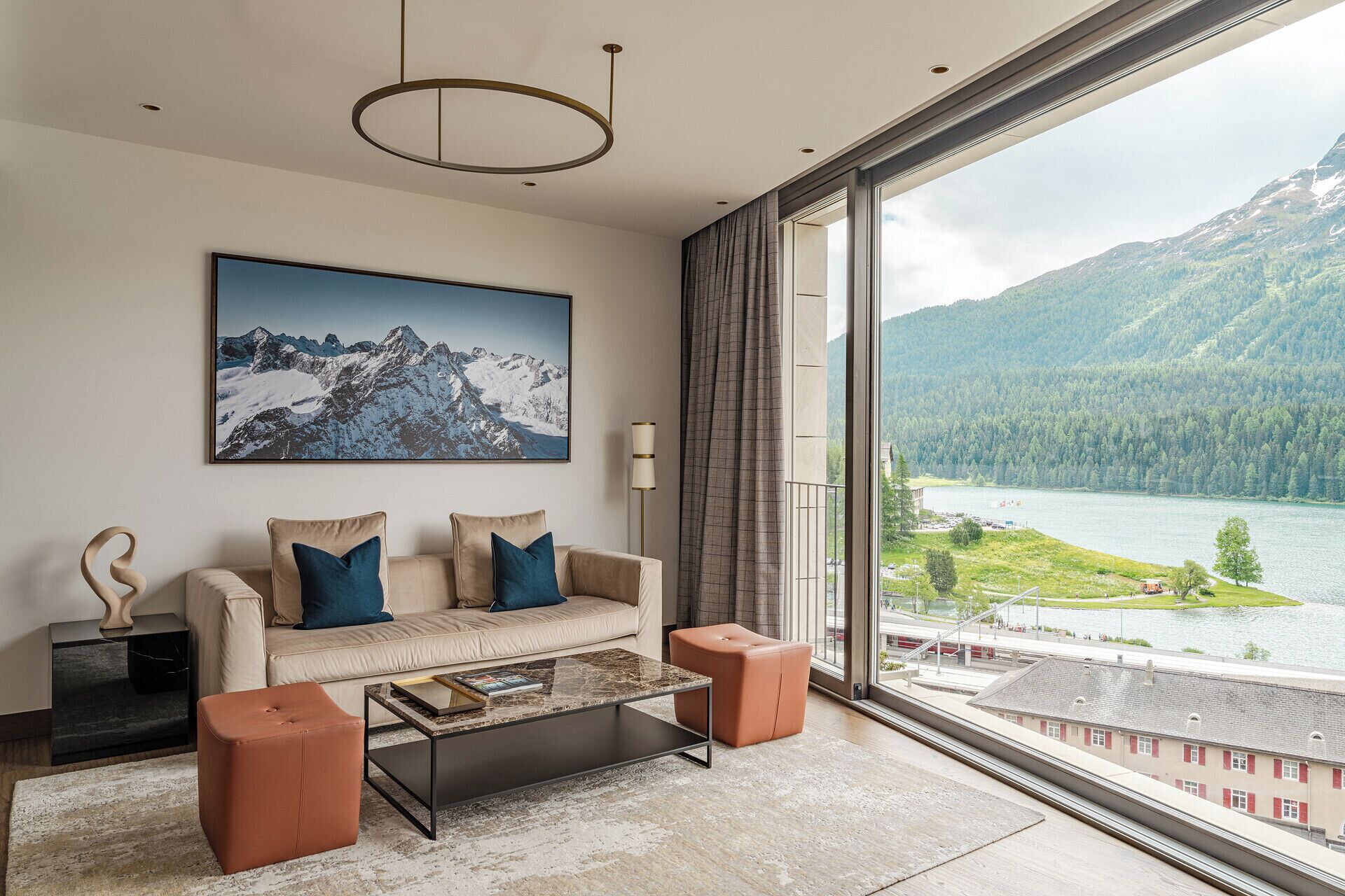 Ein modernes Wohnzimmer mit einer Couch, Hocker, einem Couchtisch und einem großen Fenster mit Blick auf einen See und Berge.