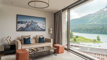 Ein modernes Wohnzimmer mit einer Couch, Hocker, einem Couchtisch und einem großen Fenster mit Blick auf einen See und Berge.