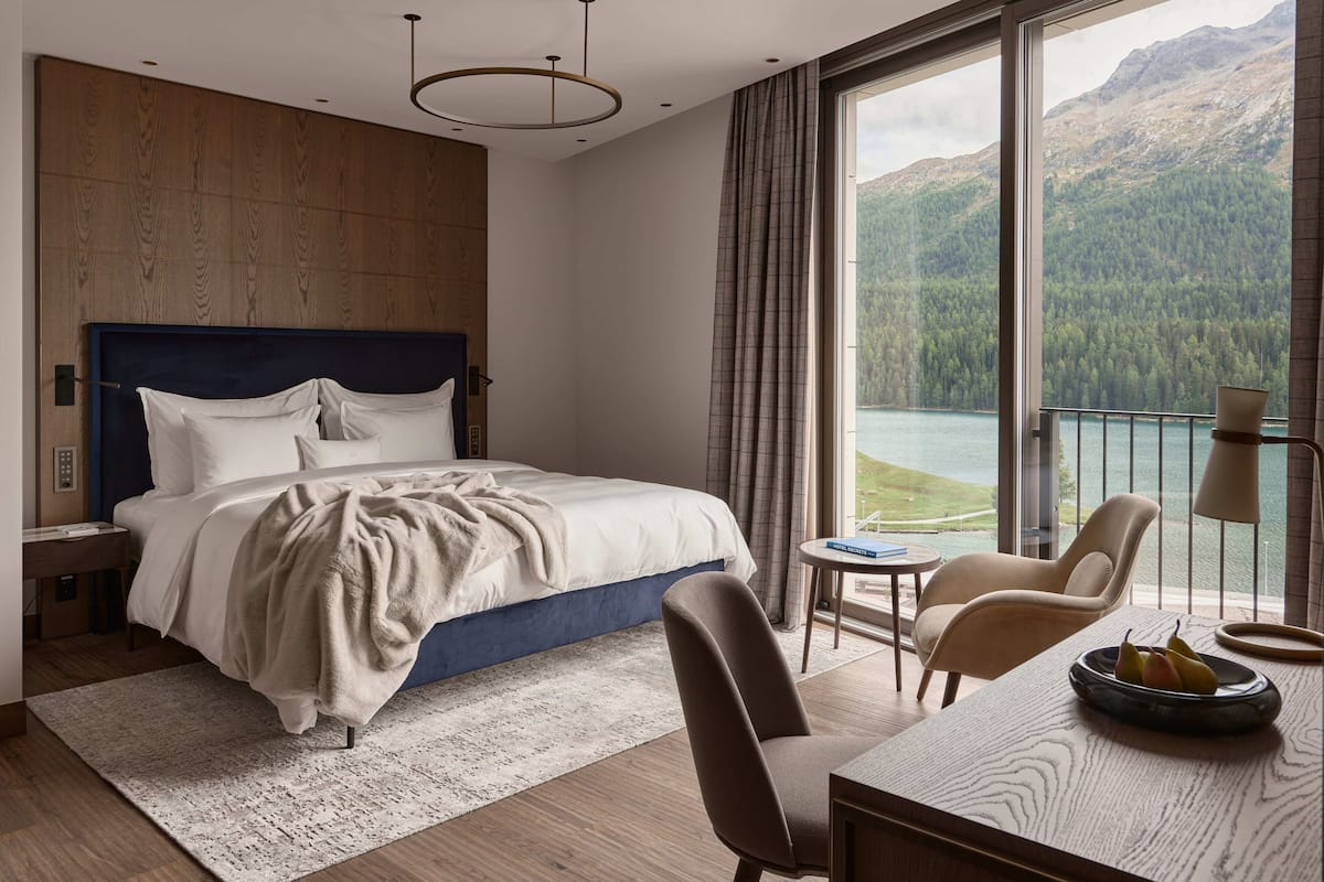 A modern bedroom with a large bed, a small table, and a chair. There is a view of mountains and a lake through the window.