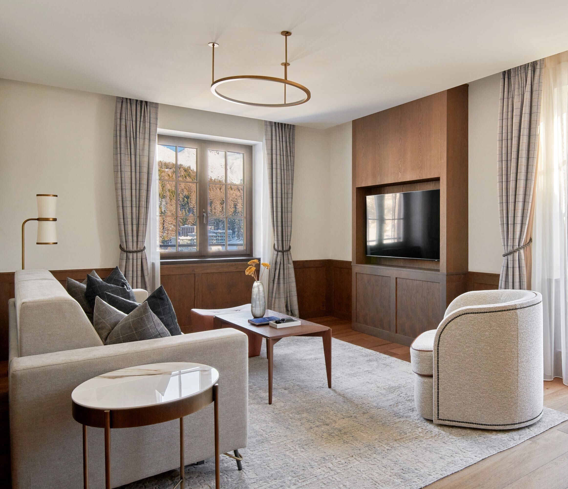 Una sala de estar moderna con sofá, sillón, mesa de centro y televisor montado en una pared de madera. Una ventana con cortinas ofrece vistas al exterior.
