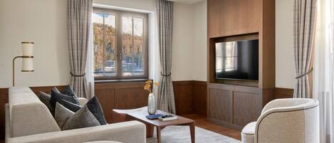 Una sala de estar moderna con sofá, sillón, mesa de centro y televisor montado en una pared de madera. Una ventana con cortinas ofrece vistas al exterior.