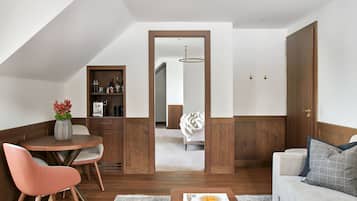 A modern living room with a round wooden table, a sofa, and a coffee table. There is a bookshelf with books and decorative items. A doorway leads to another room with a bed and a hanging light fixture.