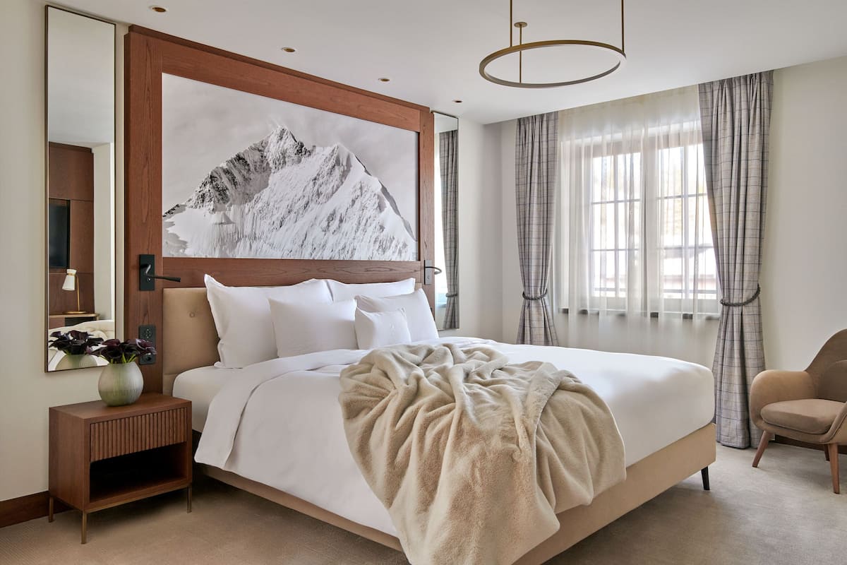 A modern hotel room with a large bed, a wooden nightstand, a mirror, and a mountain artwork on the wall.