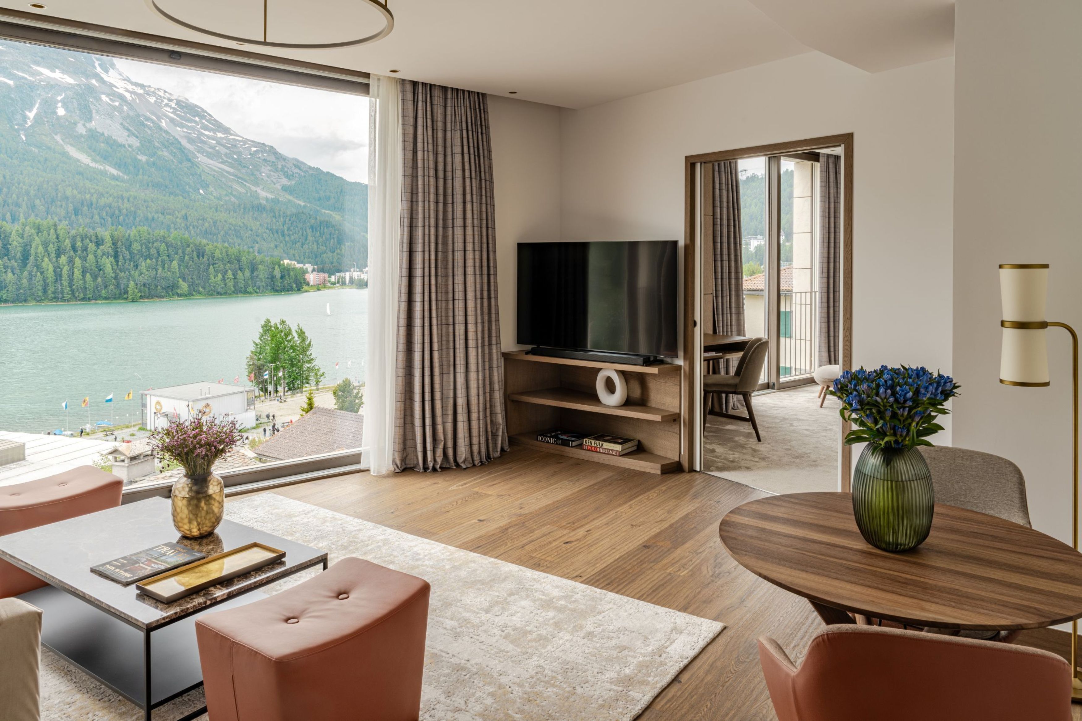 Una sala de estar moderna con un amplio ventanal con vistas a un lago y montañas, un televisor de pantalla plana y una zona de comedor con mesa y sillas de madera.