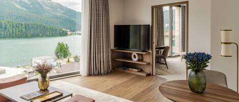 Una sala de estar moderna con un amplio ventanal con vistas a un lago y montañas, un televisor de pantalla plana y una zona de comedor con mesa y sillas de madera.