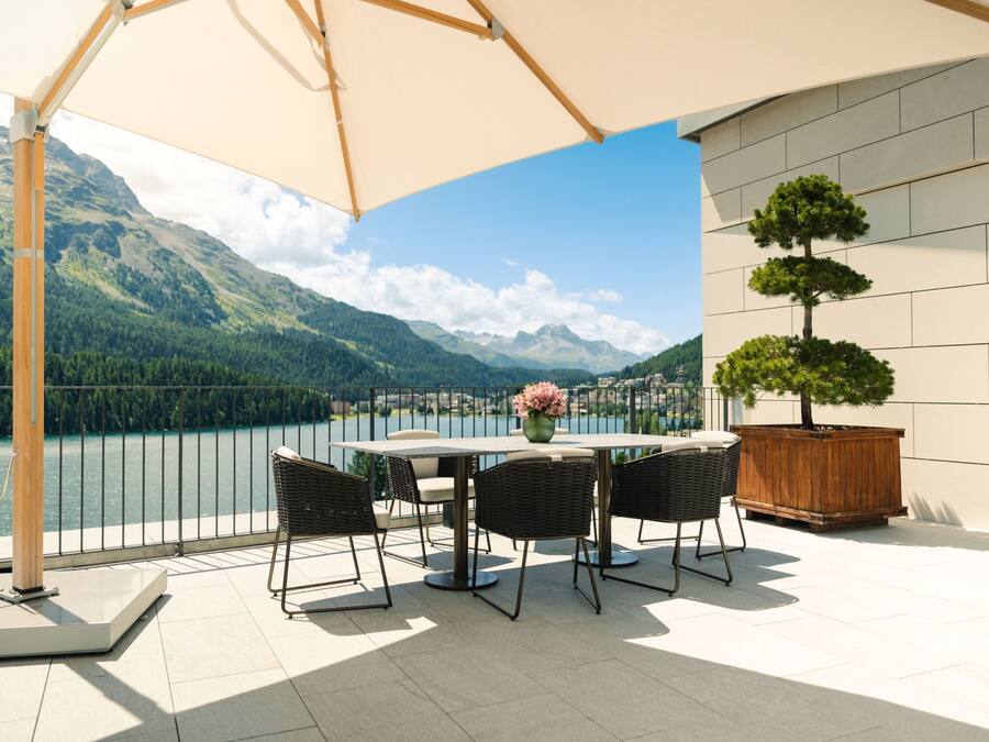 A terrace with a table and chairs, a wooden planter with a tree, and a view of mountains and a lake.
