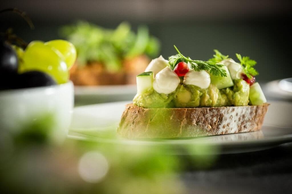Un sandwich garni d’avocat, de fromage et de légumes verts, servi sur une tranche de pain.