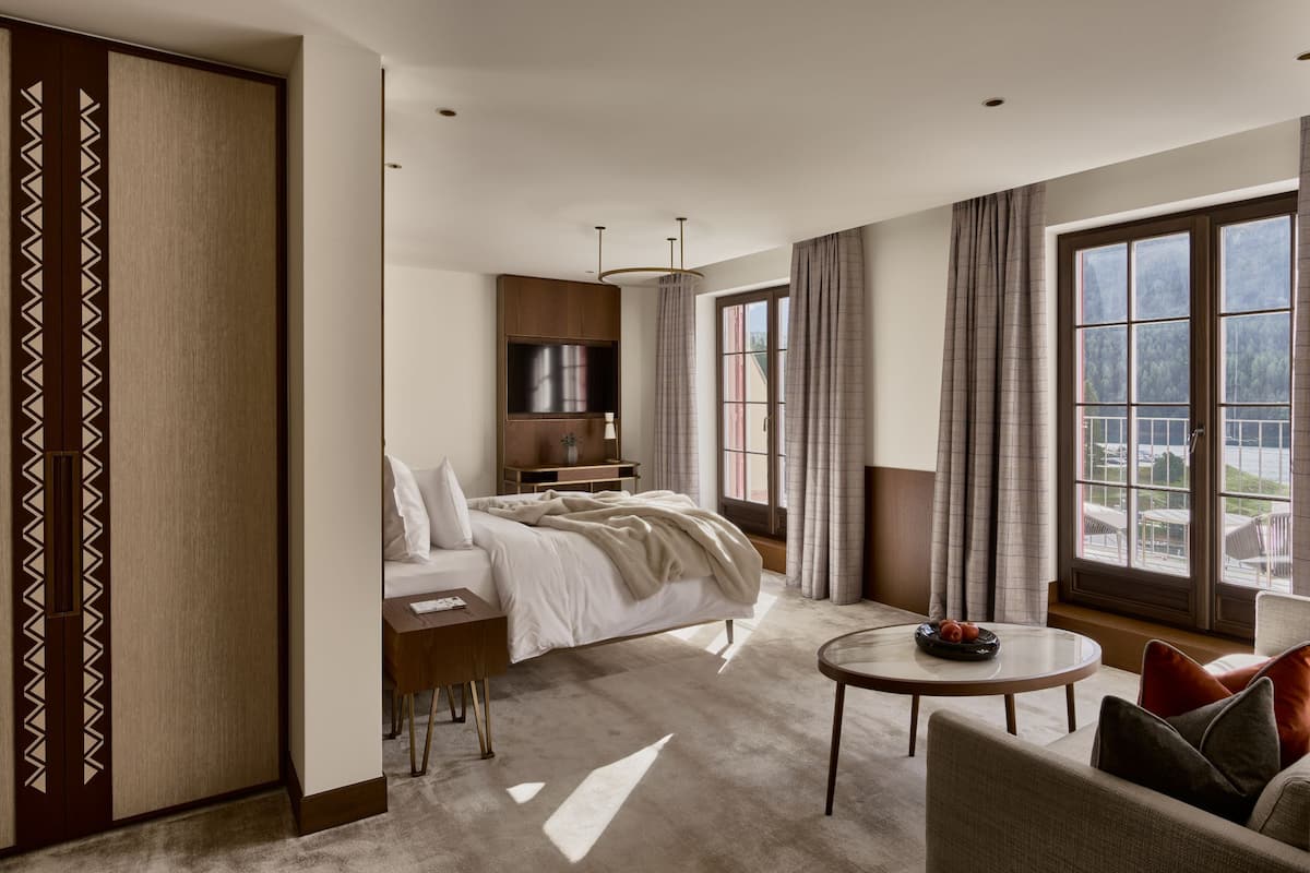 A modern hotel room with a bed, a desk, a round table, and a view of the outdoors through large windows.
