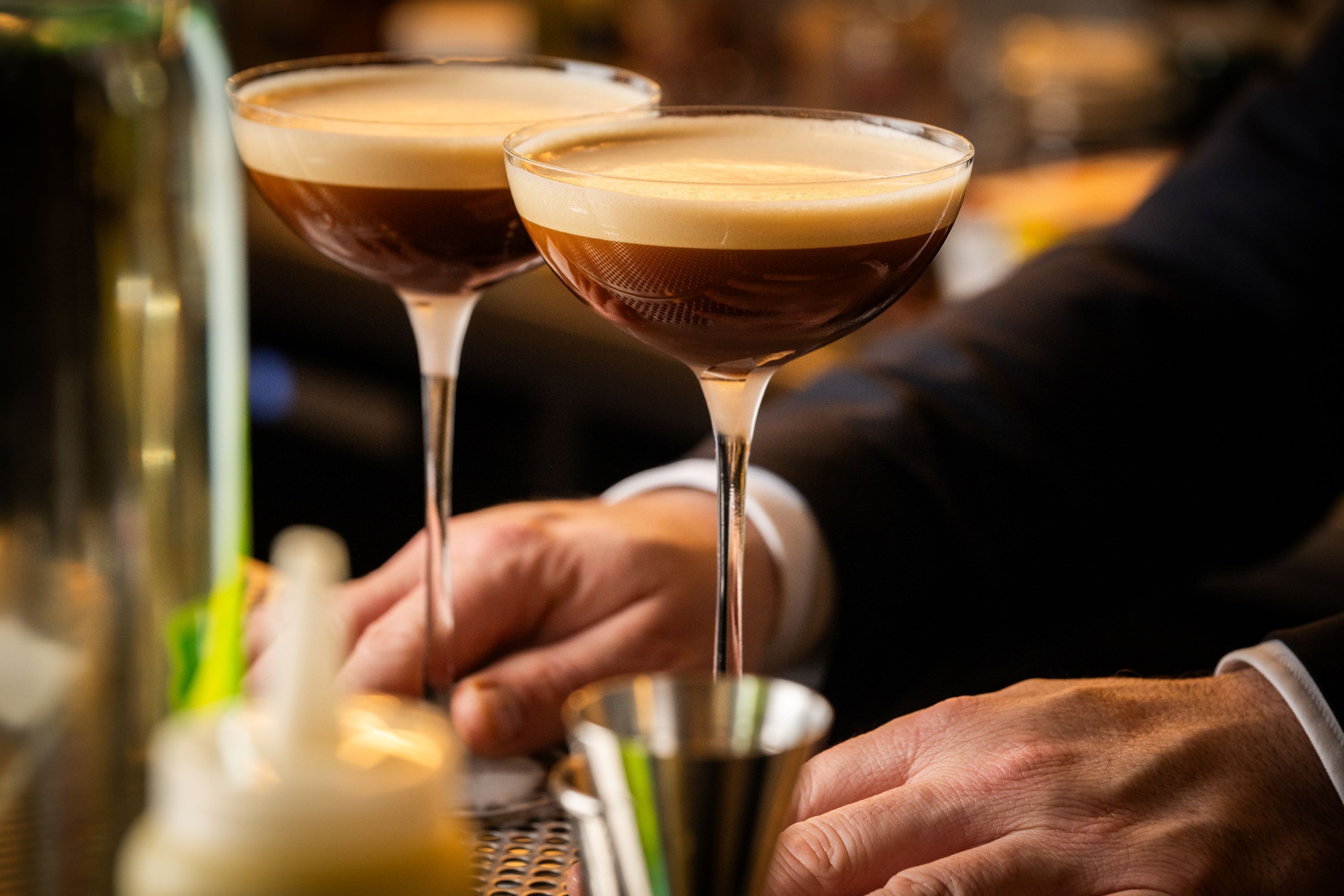 Dos copas de martini llenas de un líquido oscuro y espuma, sostenidas por una persona con traje.