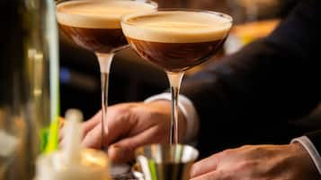 Dos copas de martini llenas de un líquido oscuro y espuma, sostenidas por una persona con traje.
