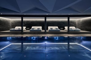 A modern indoor pool area with four reclining loungers aligned against a stone wall.