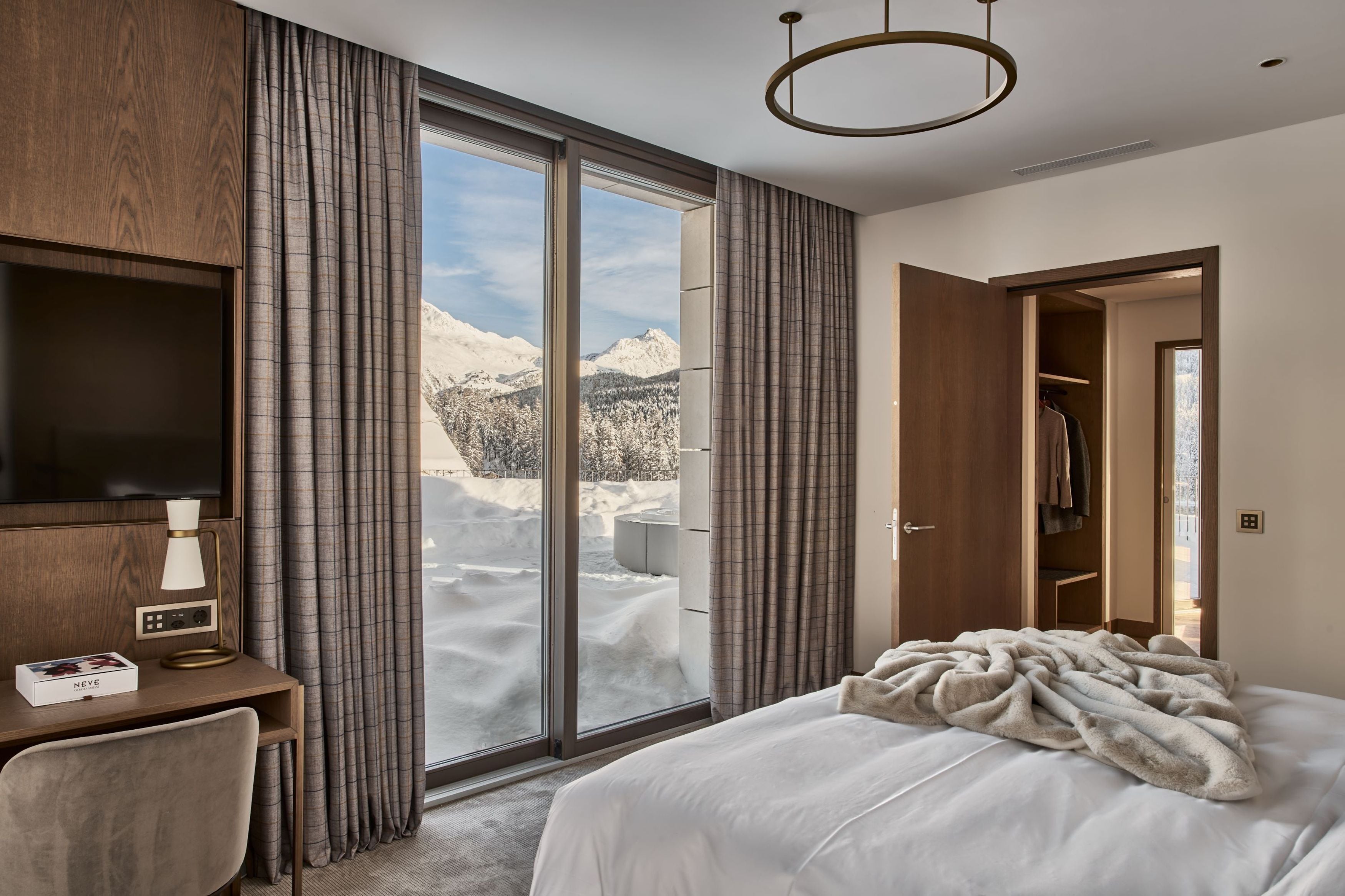 A hotel room with a large window offering a mountain view, a bed with a white comforter, a desk with a lamp, and a TV.