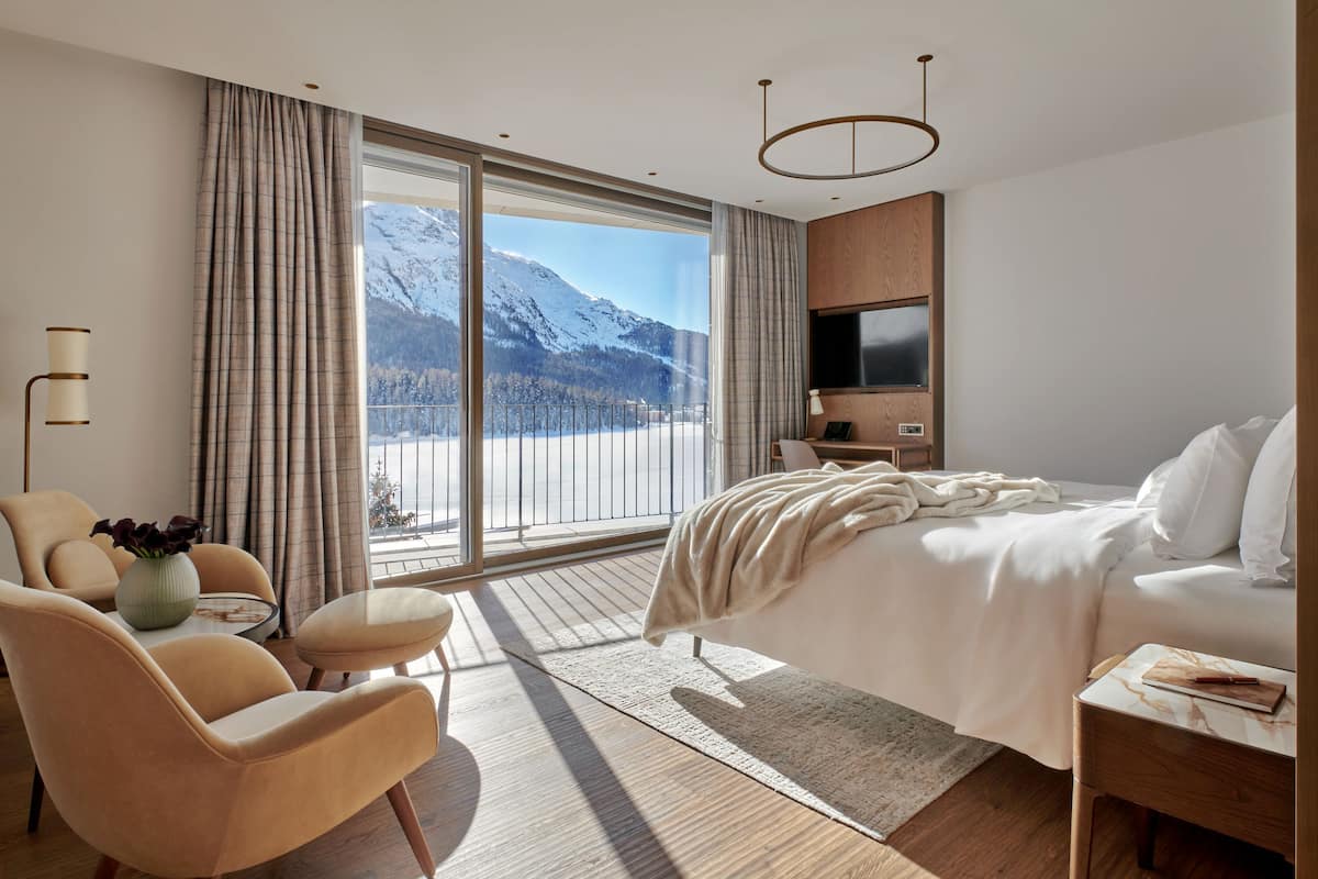 A modern hotel room with a large bed, a sofa, a TV, and a view of snow-capped mountains.