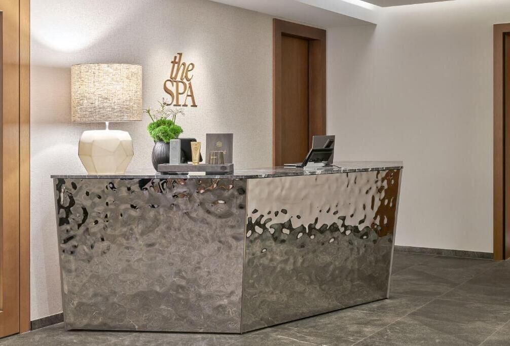 Un mostrador de recepción con superficie gris texturizada, un cartel en la pared que dice "the SPA", una lámpara, una planta en maceta y un ordenador portátil.