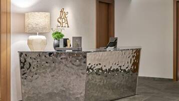 Un mostrador de recepción con superficie gris texturizada, un cartel en la pared que dice "the SPA", una lámpara, una planta en maceta y un ordenador portátil.