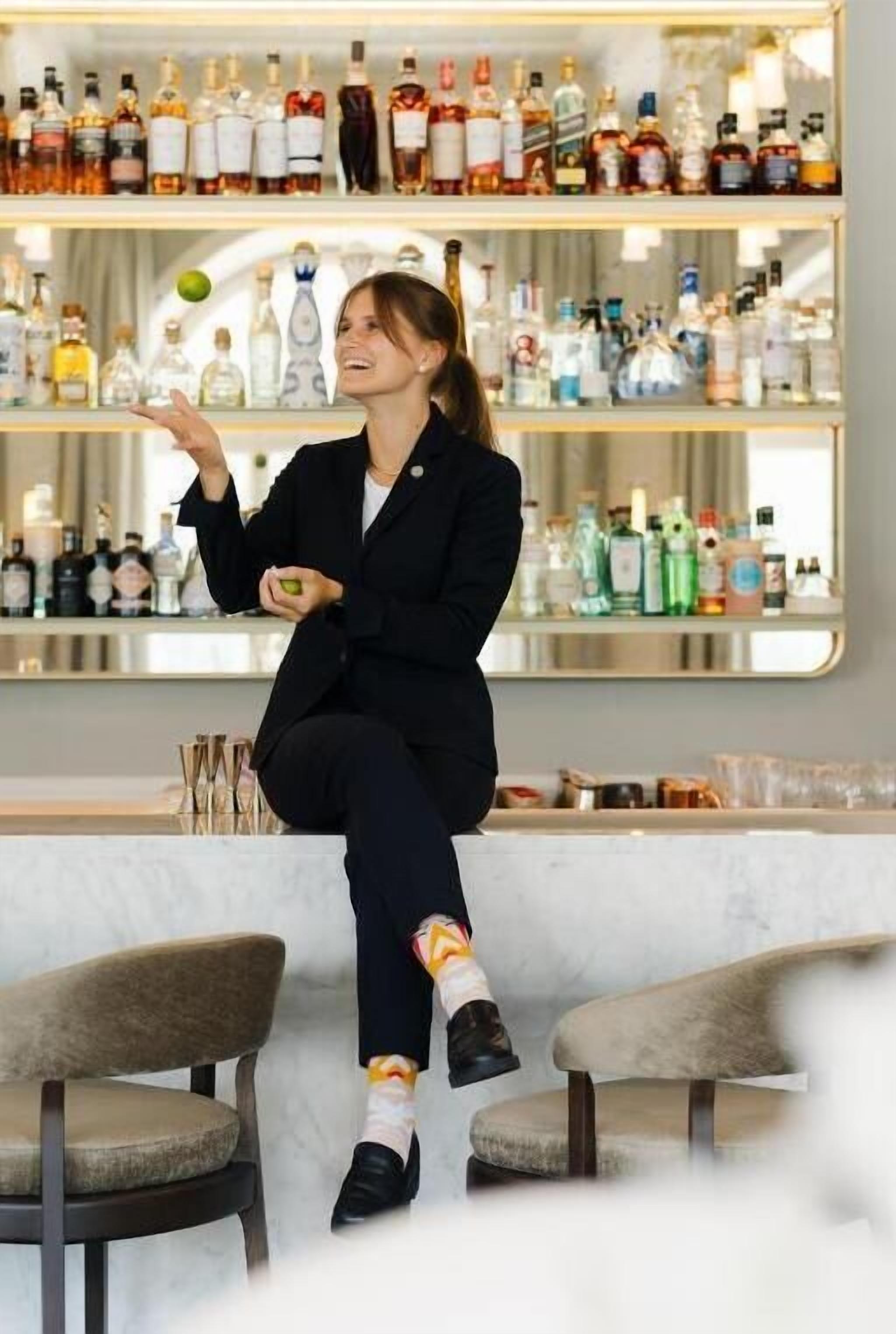 Una persona seduta su uno sgabello da bar in una stanza con scaffali pieni di bottiglie di liquore sullo sfondo.