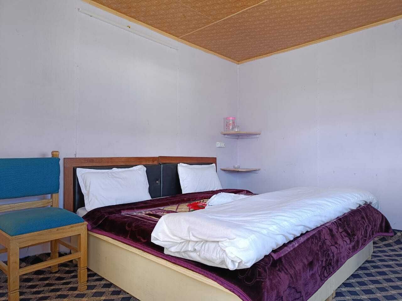 A single bed with a wooden headboard, a blue upholstered chair, and a small shelf with a pink object.